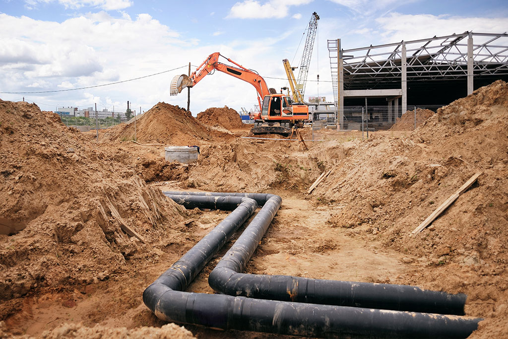 Construction site featuring heavy machinery, including an excavator and cranes, with exposed piping and mounds of dirt, illustrating Clearwater Plumbing and Civil's civil works services in Sydney.
