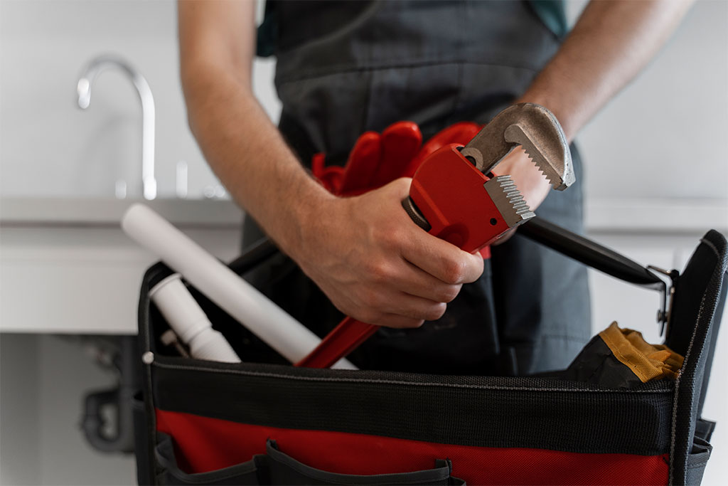 Plumber holding a red adjustable wrench above a tool bag containing plumbing supplies, with a kitchen sink in the background, representing Clearwater Plumbing and Civil's professional plumbing services.