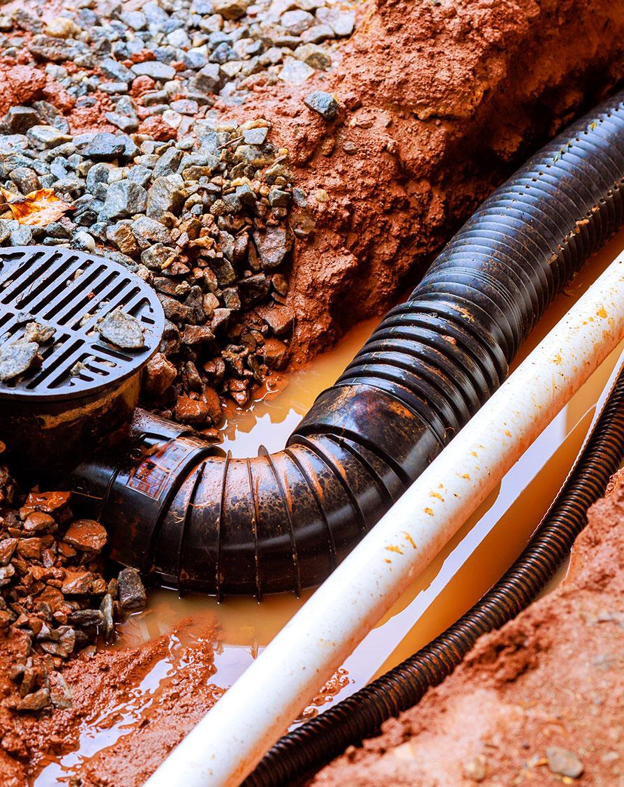 Drainage system with visible water pooling, black drainage pipe, and gravel, illustrating plumbing issues relevant to emergency repair services.